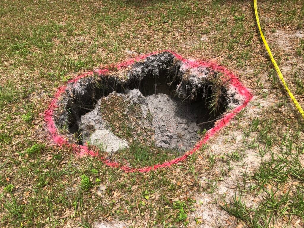 Circular sinkhole in a grassy lawn marked with pink spray paint and yellow caution tape